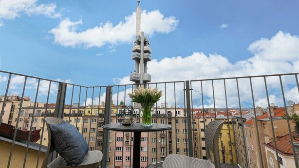Ein Balkon mit Tisch, zwei Stühlen und einem Blumenarrangement. Im Hintergrund ist ein hoher Turm sichtbar.