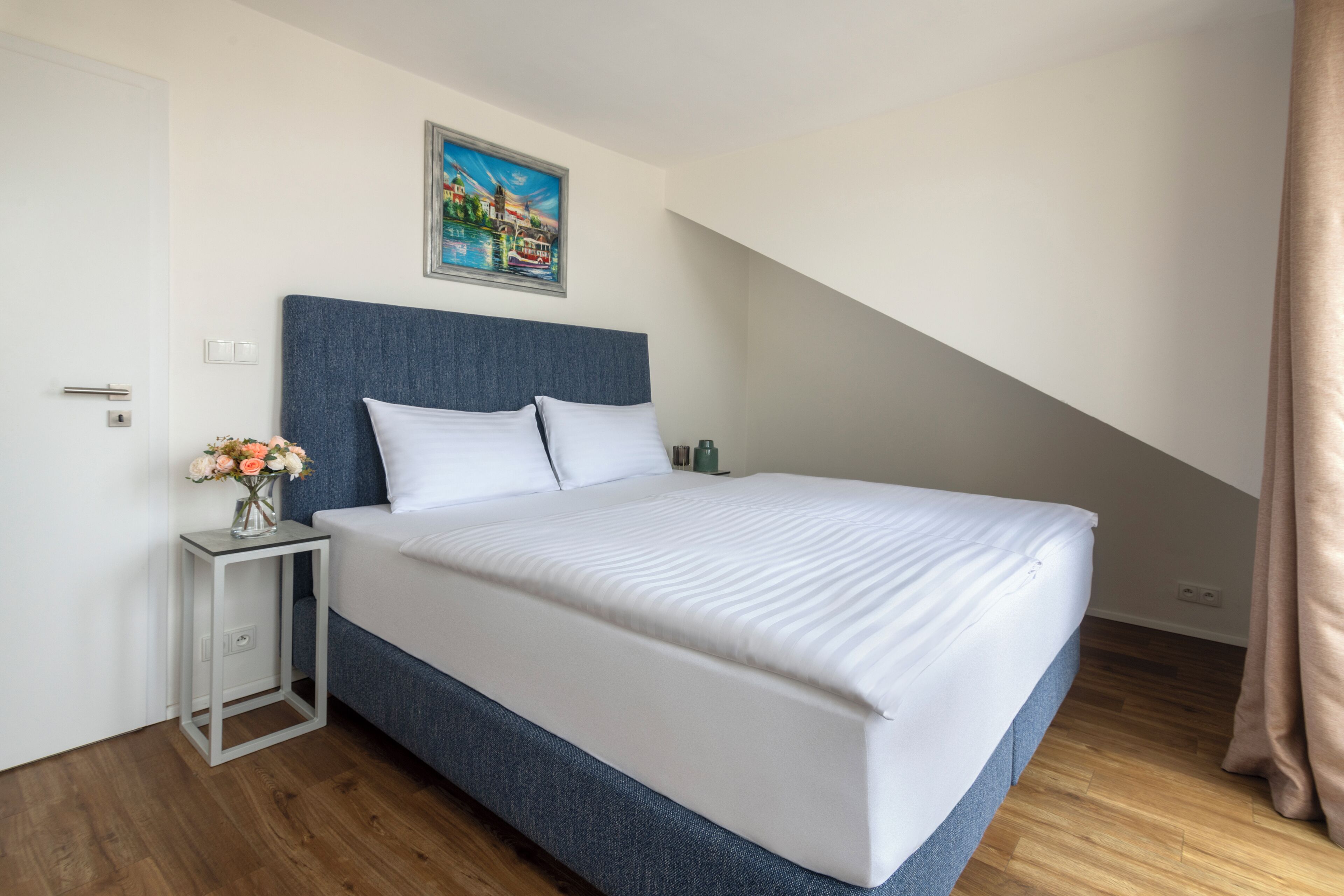 A modern hotel room with a large bed, a small bedside table with a vase of flowers, a painting on the wall, and a wooden floor.