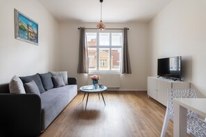 Een moderne woonkamer met een grijze bank, een klein rond tafeltje met een bloevas, een wit kastje met een flatscreen televisie en een raam met gordijnen.