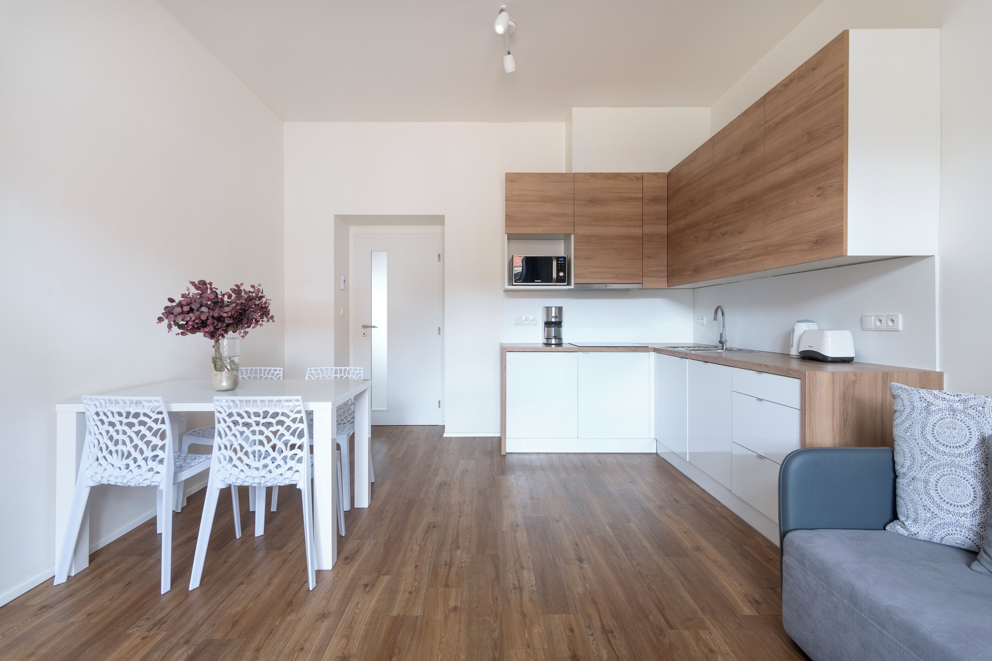A modern kitchen with white cabinetry, a microwave, and a coffee maker. There is a dining table with white chairs and a vase with flowers.