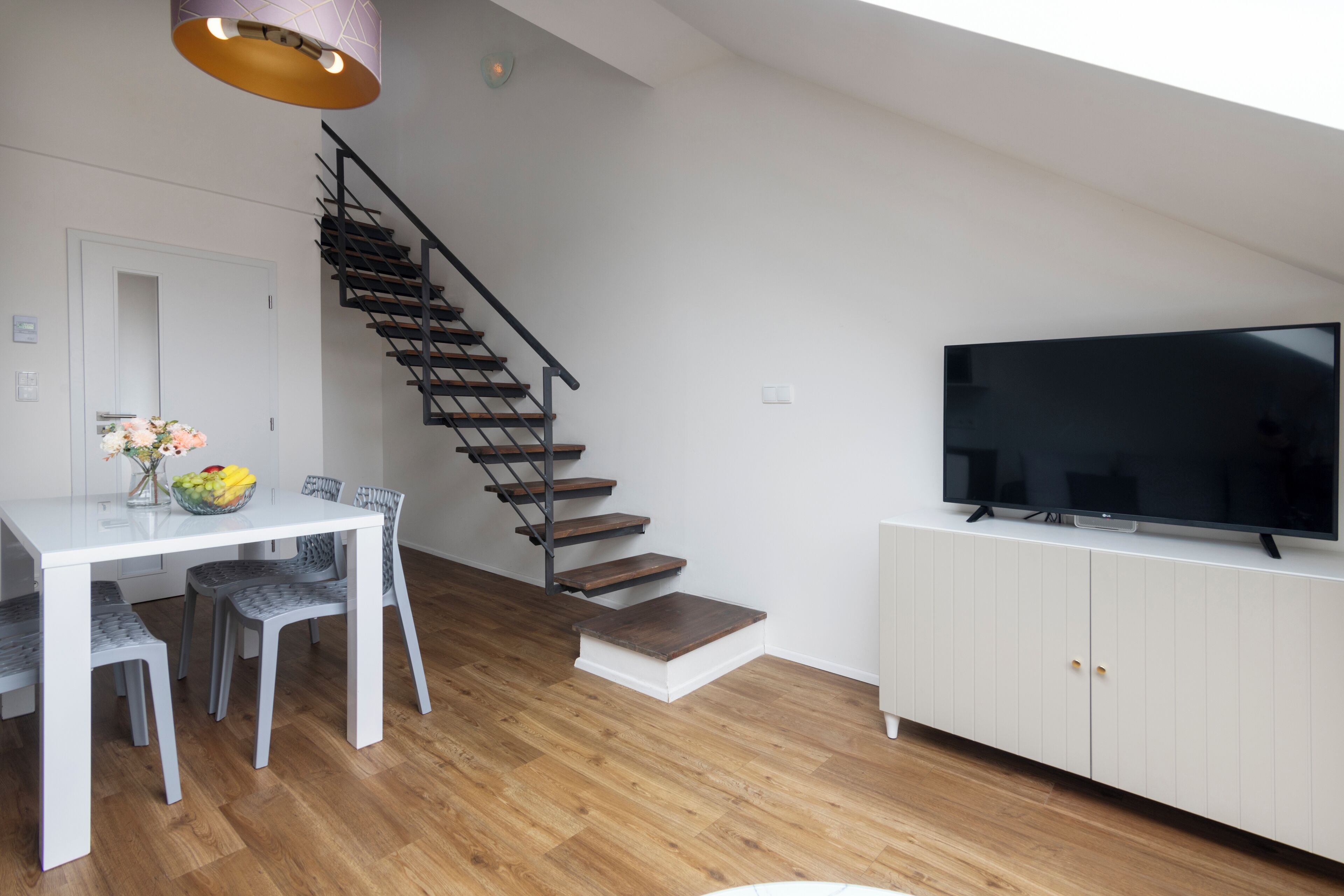 Una sala de estar moderna con una escalera, un comedor con una mesa y sillas blancas, y un televisor de pantalla plana sobre un mueble blanco.