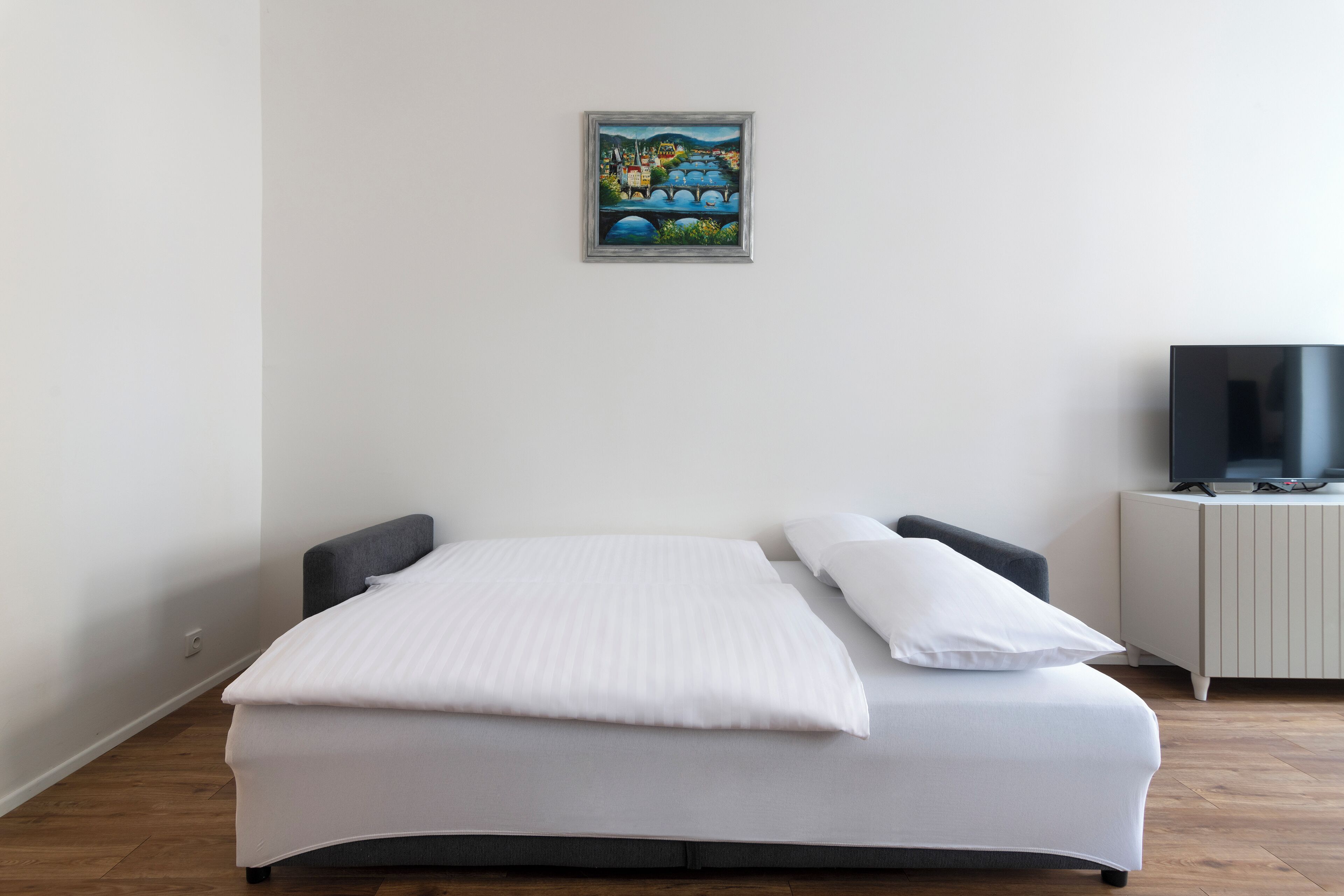 Een net opgemaakt bed met witte lakens, een grijs hoofdbord en één kussen. Daarboven hangt een lijst met een kunstwerk aan de muur.