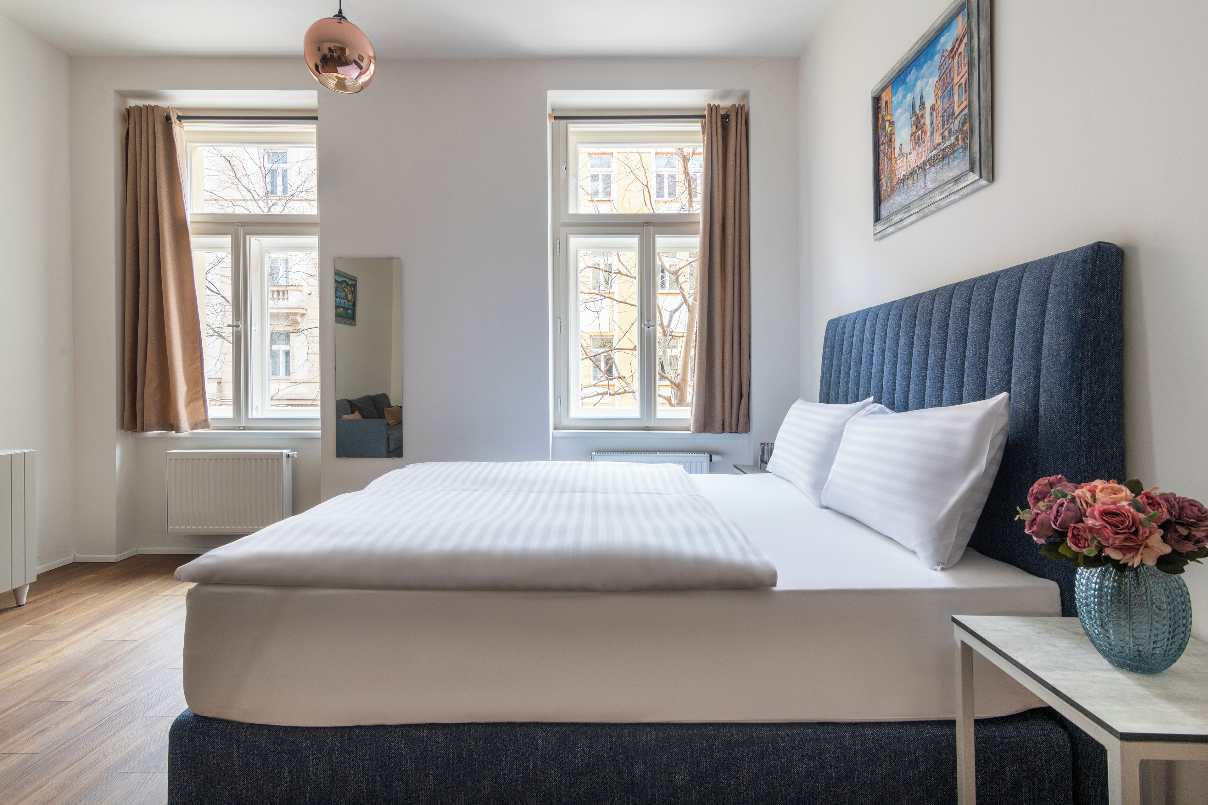 Habitación de hotel con una cama grande, dos ventanas con cortinas, un radiador, una mesita de noche con un jarrón de flores y un cuadro en la pared.