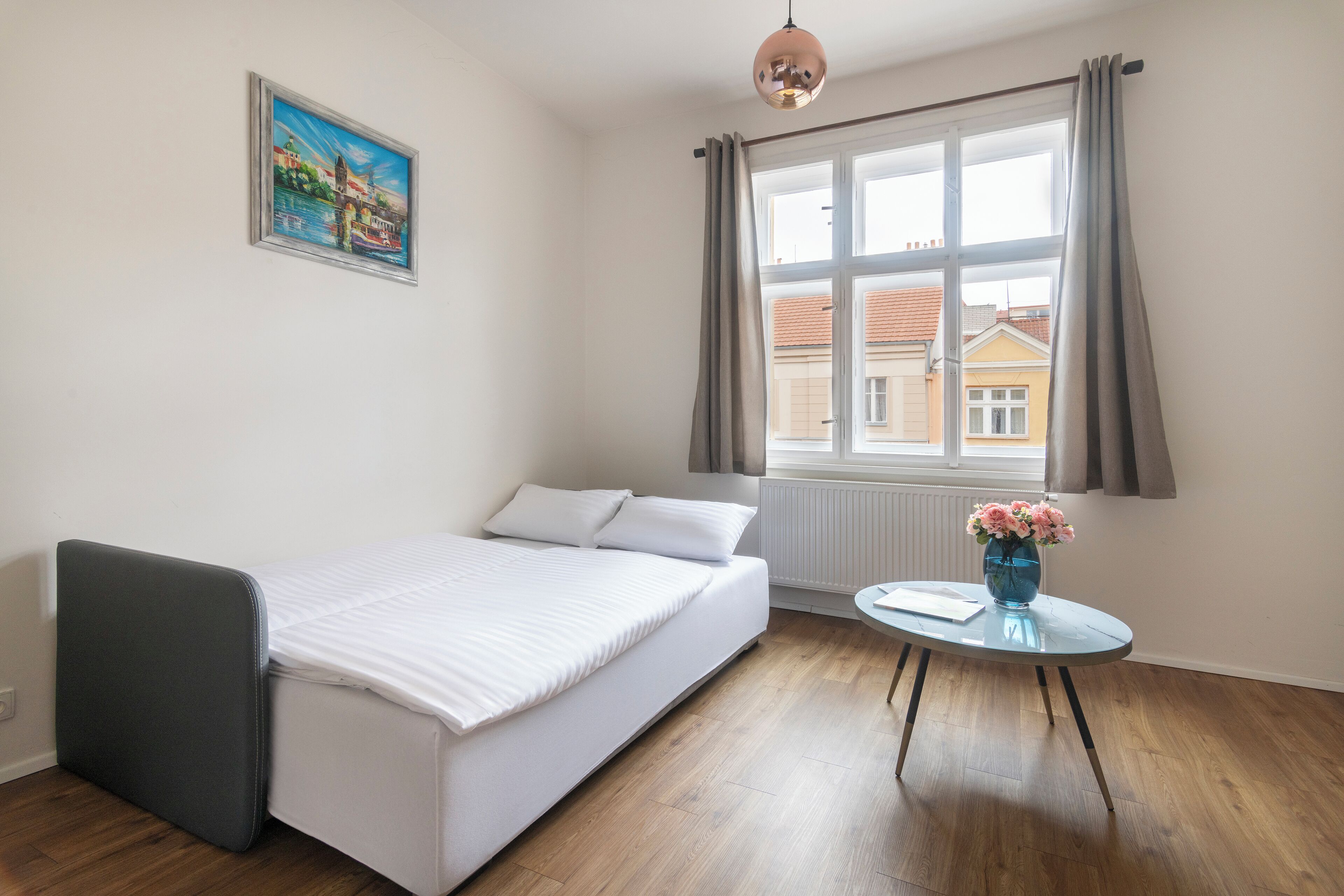 A room with a bed, a small table with a vase, a window with curtains, and a painting on the wall.