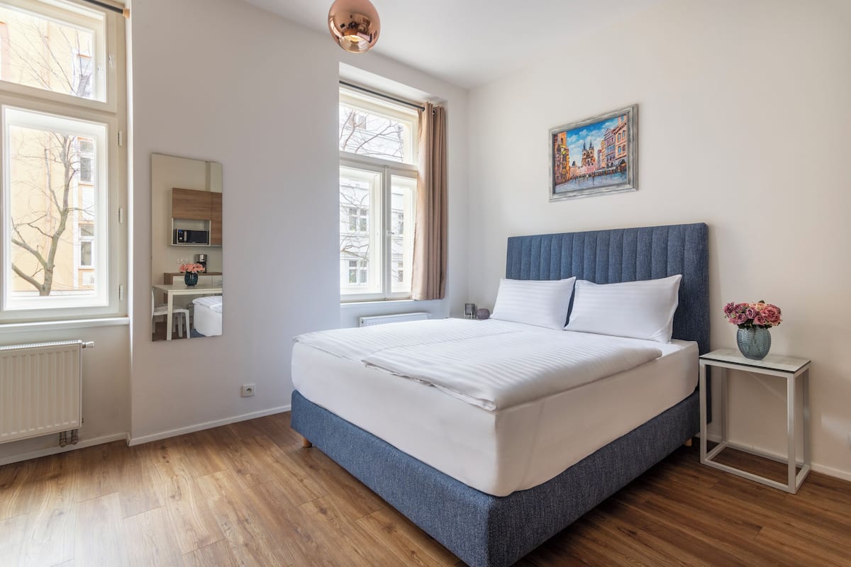 Une chambre avec un grand lit, une table de chevet ornée d’un vase, un tableau accroché au mur et une fenêtre donnant sur des immeubles.
