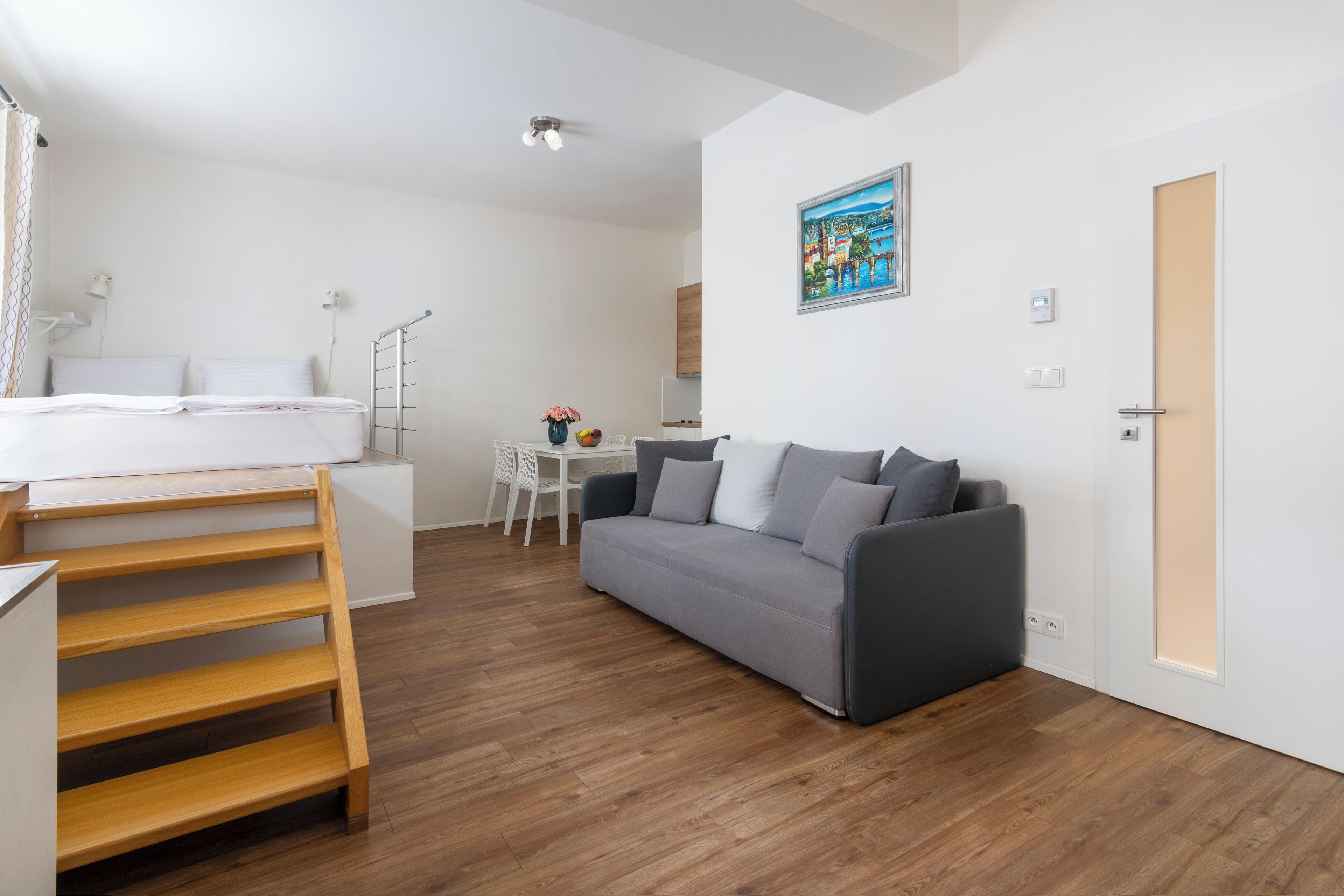 Una habitación moderna y luminosa con piso de madera, un sofá gris, una mesita blanca pequeña y una cama con escalera de madera.