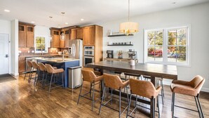A modern kitchen with wooden cabinets, a central island with stools, a large dining table, and a hanging light fixture. - Sister Lakes 7 bedroom with Game room, Hot tub, Basketball and amazing views (Dowagiac)