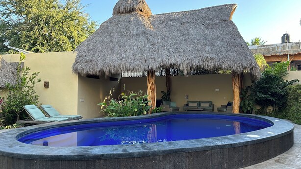 Una piscina con una estructura de techo de paja, sillas de descanso y un jardín tropical.