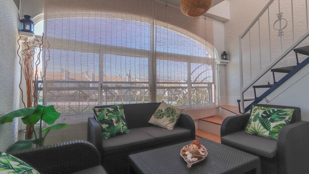 A modern living room with a large window, a staircase, and a wicker sofa with green cushions. - 090 - Altomar II 005 - comfortHOLIDAYS (el Gran Alacant)
