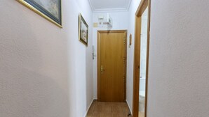 Un pasillo estrecho con una puerta de madera al final, paredes blancas y suelo de baldosas.