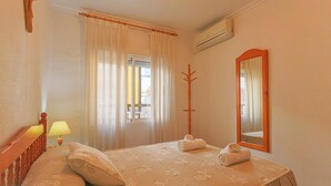 Una habitación de hotel con cama, mesita de noche con lámpara, ventana con cortinas, perchero de madera y espejo.