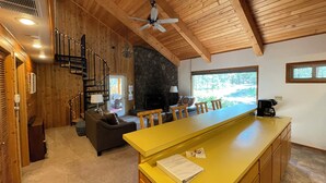 A spacious living area with a wooden ceiling, a stone fireplace, a dining area with a yellow counter, and a view of the outdoors.
