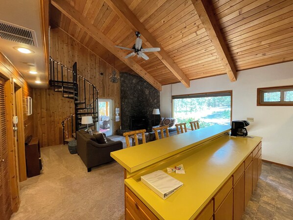 Uma sala de estar ampla com teto de madeira, lareira de pedra, área de refeições com balcão amarelo e vista para o exterior.