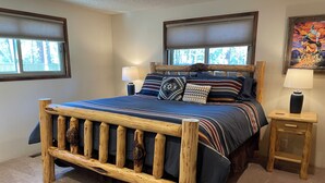 A bedroom with a wooden bed frame, a large window with blinds, a nightstand, and a lamp.