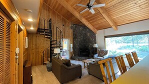 A cozy cabin interior with a stone fireplace, wooden walls, a spiral staircase, and a dining area with wooden chairs.