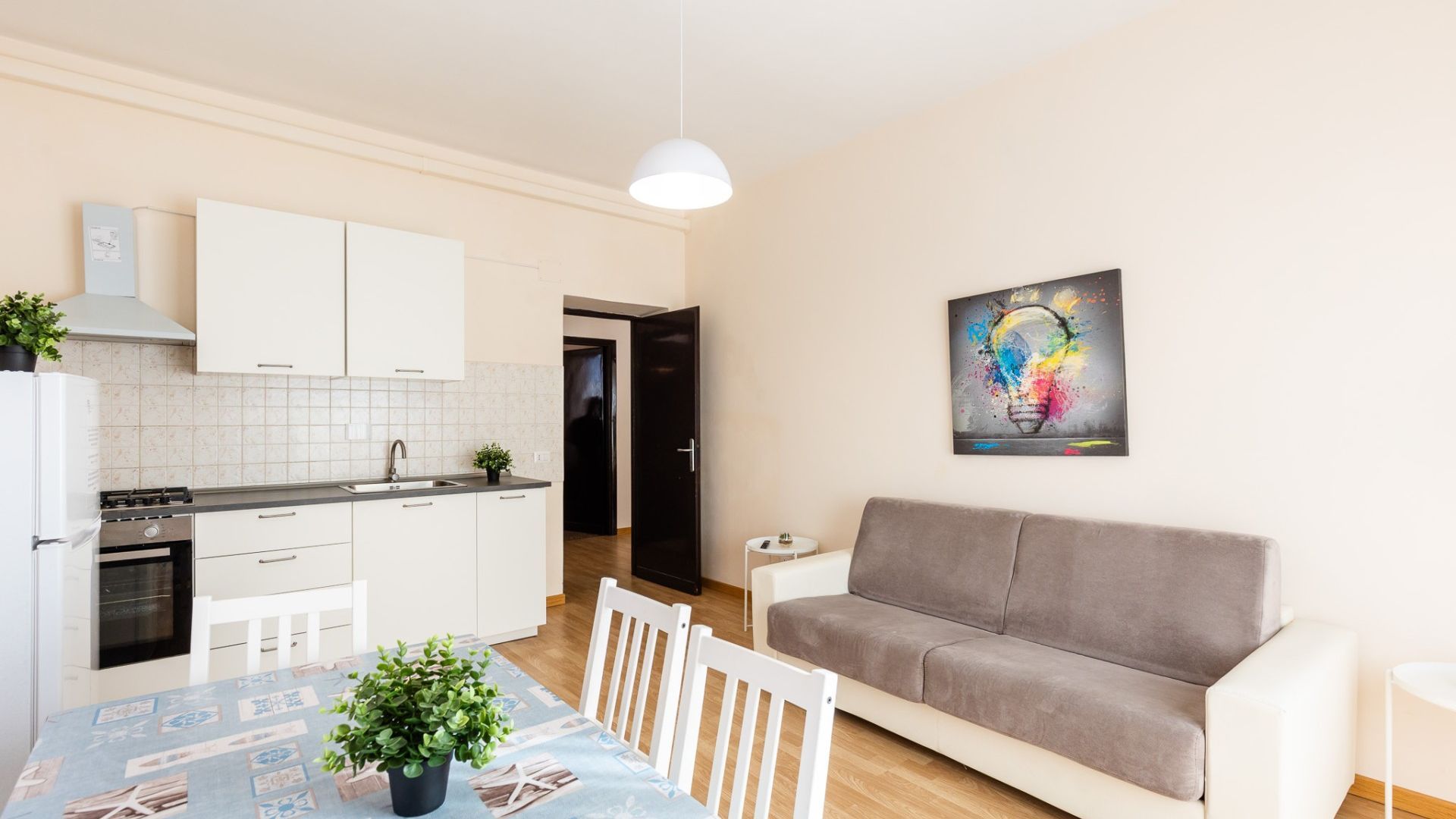 Une cuisine moderne avec des meubles blancs, une table à manger avec des chaises, un canapé gris et une œuvre d’art murale représentant une ampoule colorée.
