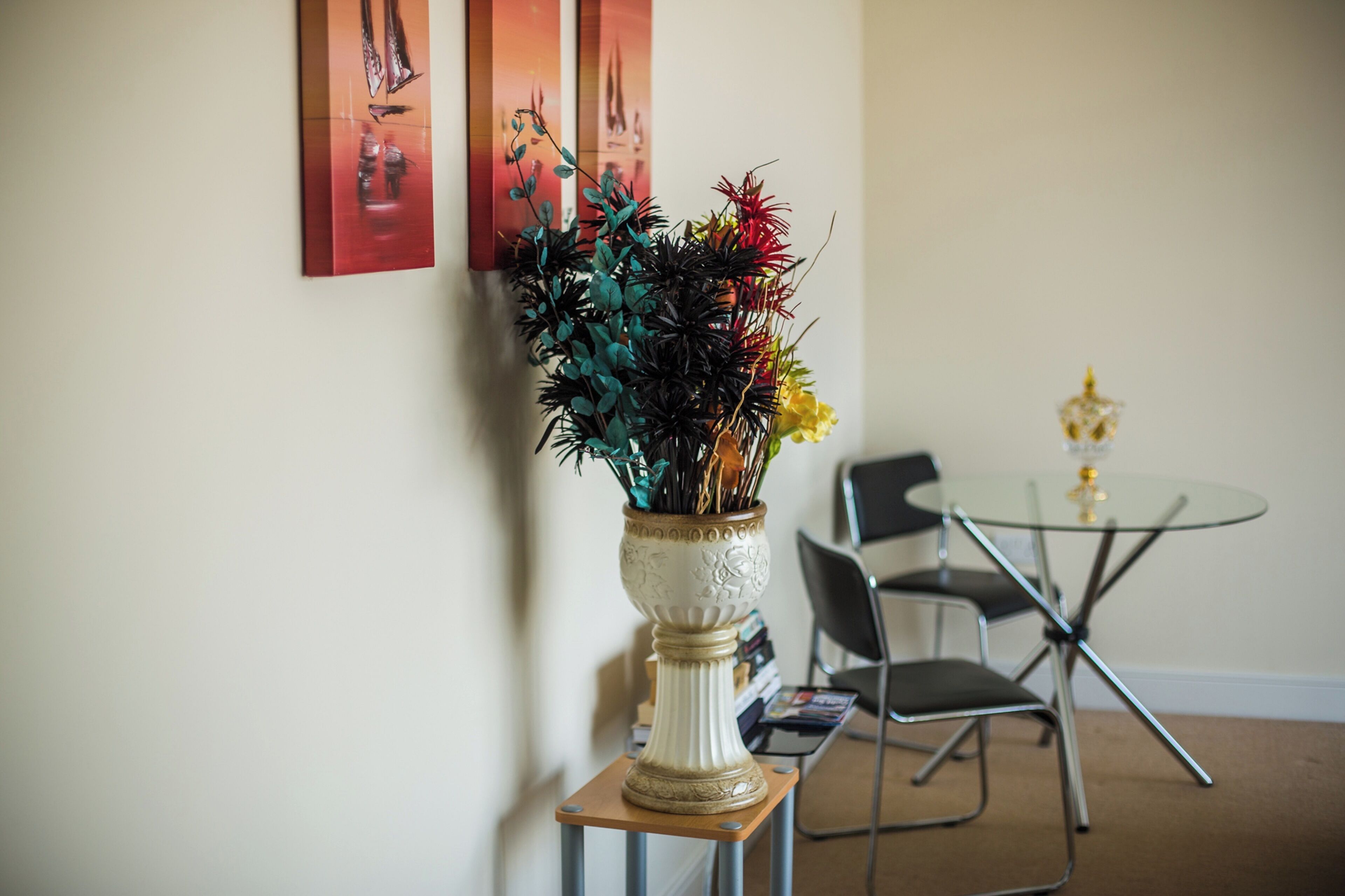 Een kamer met een witte vaas met groene en rode planten op een houten tafel, een glazen tafel met stoelen, en drie schilderijen aan de muur.