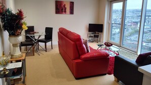 Una sala de estar moderna con un sofá rojo, una mesa de centro de vidrio, un televisor sobre un mueble bajo y vistas a la ciudad a través de amplios ventanales.
