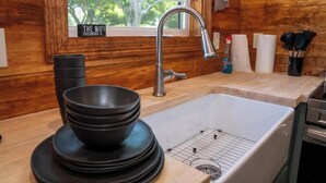A kitchen with a wooden interior, a stainless steel sink, and a window with a view of trees. - Deluxe Rustic Tiny Cabin (Broussard)