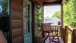 A wooden porch with a rocking chair, a door, and a view of a white RV in the background. - Deluxe Rustic Tiny Cabin (Broussard)
