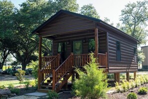Une cabane en bois dotée d’un porche et d’un escalier, entourée d’arbres et d’arbustes.