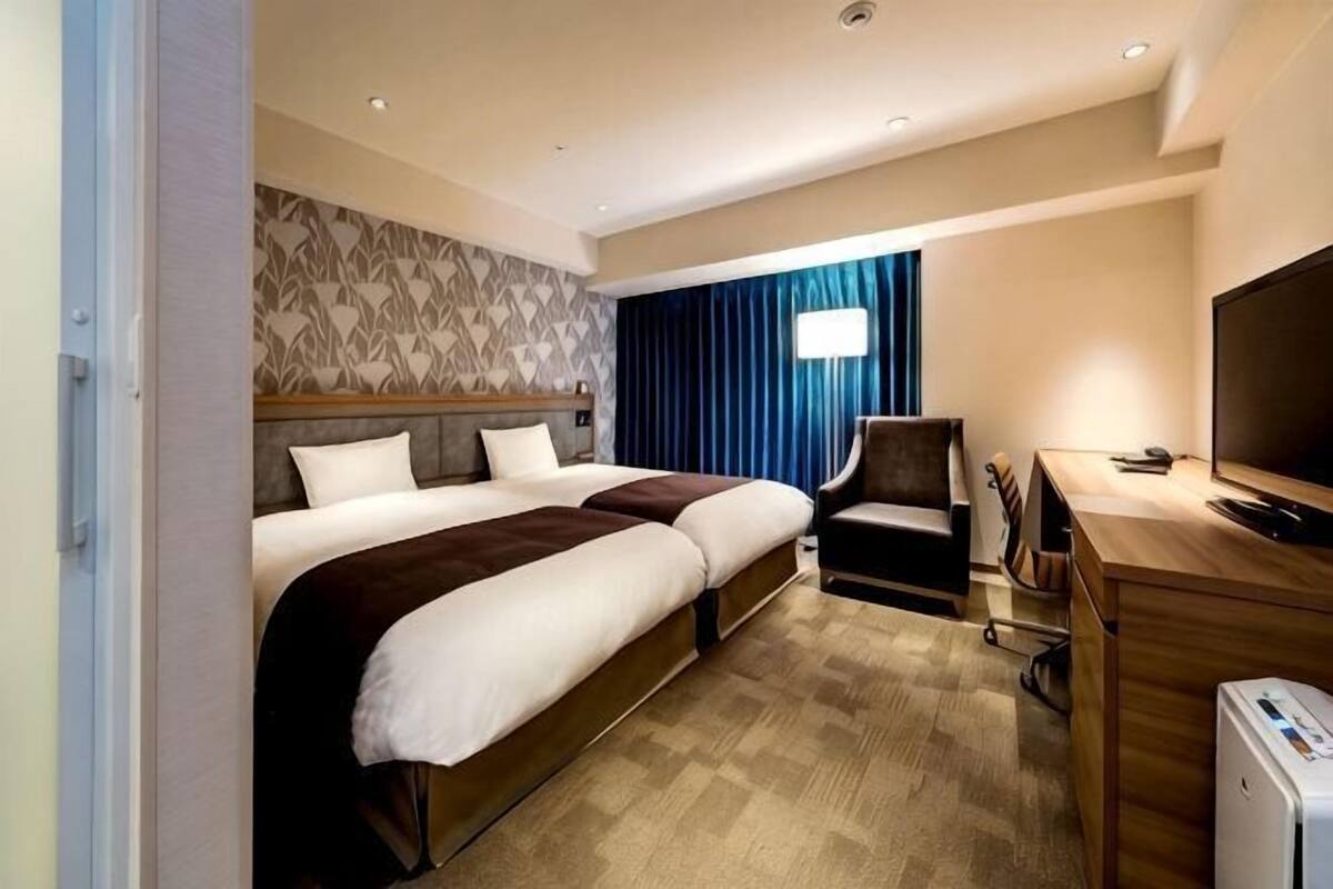 A hotel room with two beds, a desk, a television, and a window with curtains.