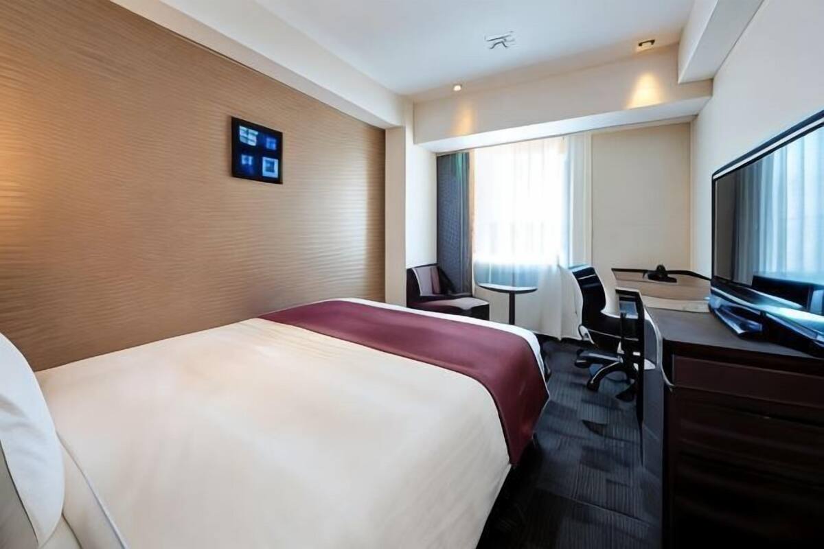 A hotel room with a bed, desk, chair, TV, and a window with curtains.