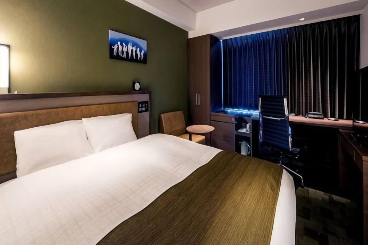 A hotel room with a bed, a desk, a chair, a small table, a TV, and a window with curtains.