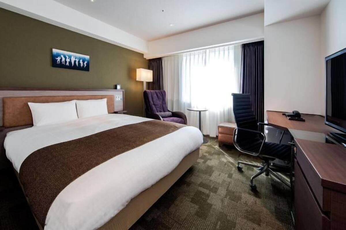 A hotel room with a large bed, a desk with a chair, a television, and a window with curtains.