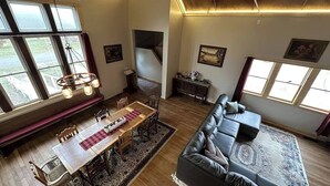 A spacious living room with a large black leather sofa, a wooden dining table with chairs, and a bench. There are multiple windows providing natural light and a view of the outdoors. - Sanctuary Parish 18Ft ceilings, 5 bedrooms, & 3 baths (Danbury)