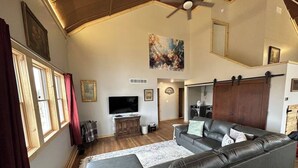 A spacious living room with a large leather sofa, a flat-screen TV, wooden flooring, and a ceiling fan. - Sanctuary Parish 18Ft ceilings, 5 bedrooms, & 3 baths (Danbury)