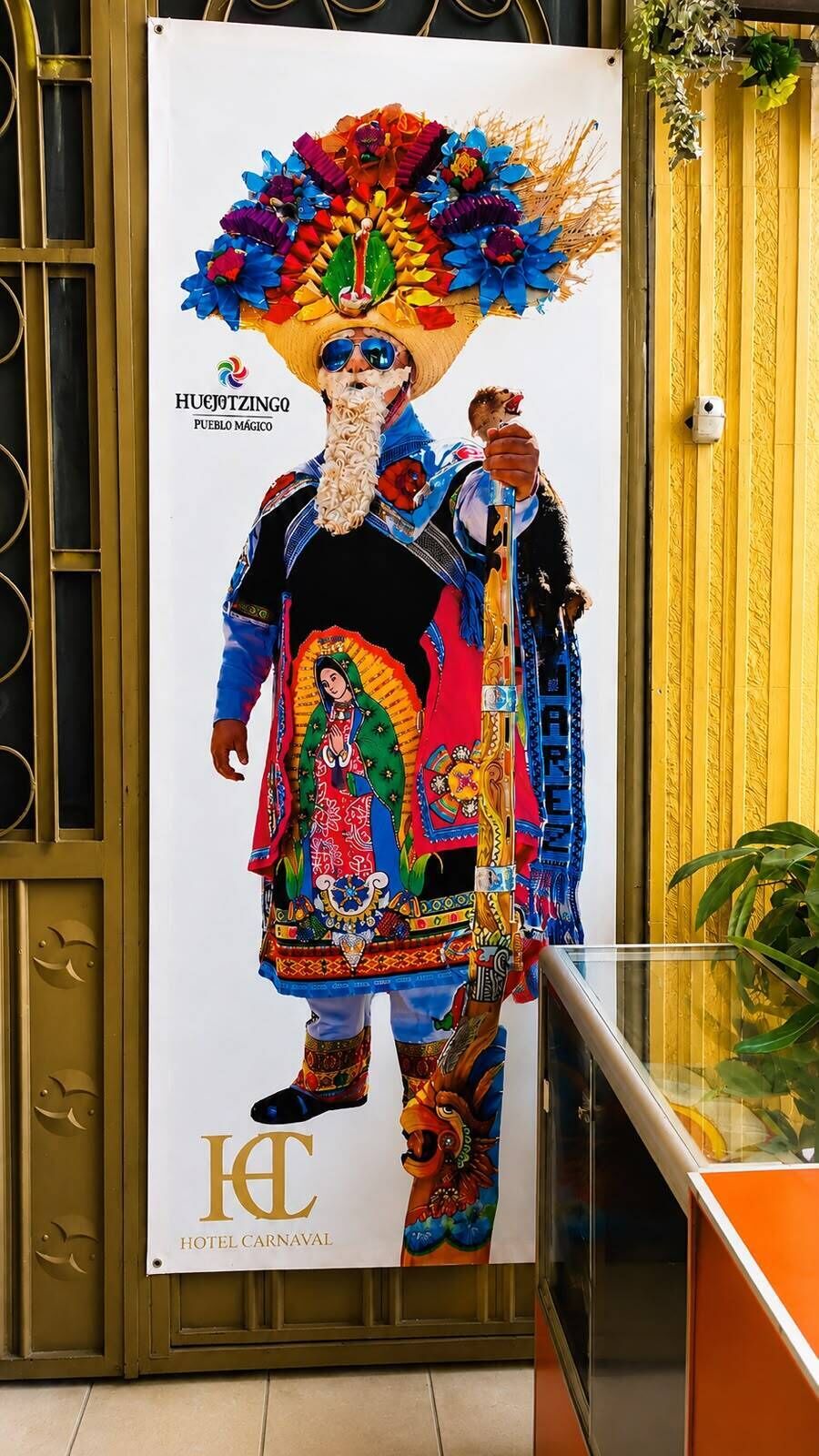Un cartel con una figura colorida, el texto 'HUEJOTZINGO PUEBLO MAGICO' y 'HOTEL CARNAVAL'.
