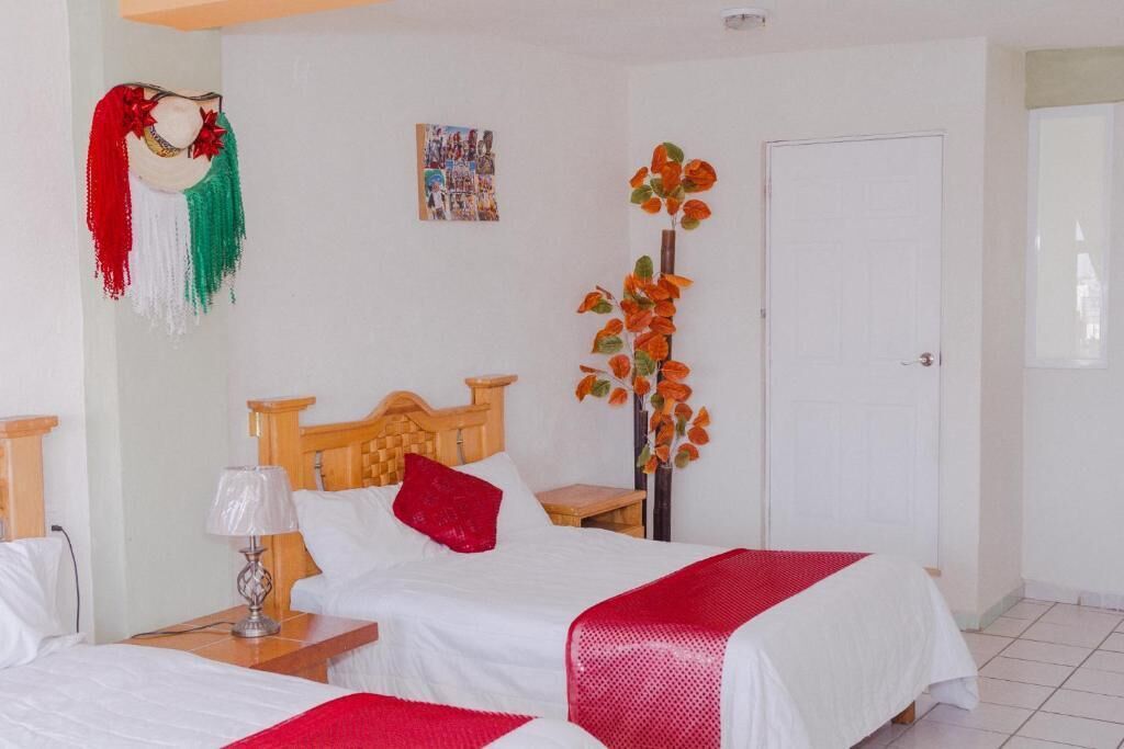 Habitación de hotel con dos camas, cabecero de madera, mesita de noche con lámpara, manta roja y una planta colgante en la pared, además de una puerta.