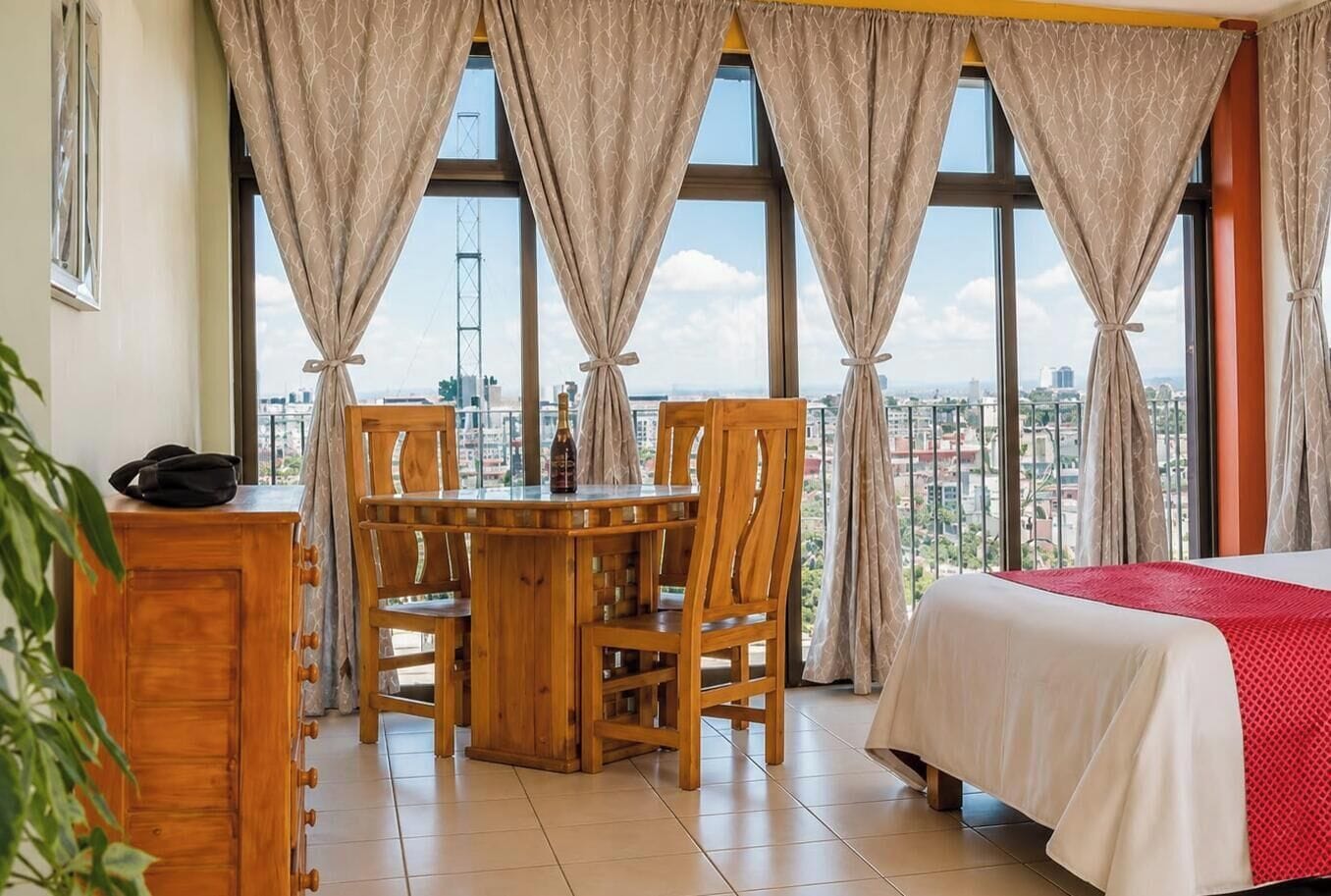 A hotel room with a bed, a dining table with chairs, a small desk, and a view of the city through large windows.