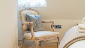 Una stanza con una poltrona classica vicino a una finestra, un comodino e un letto con copriletto bianco.