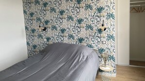 A bedroom with a bed, a wall with tropical wallpaper, a hanging light fixture, and a closet. - Logement Une Petite Escale Lectoure (Lectoure)