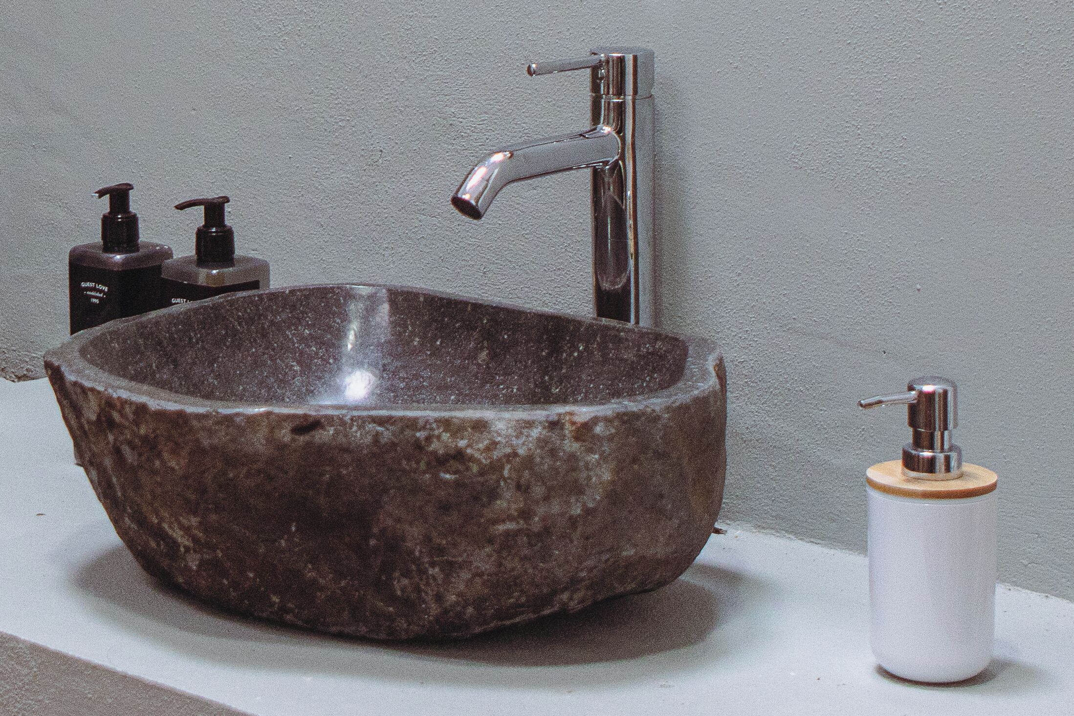 A stone sink with a modern faucet, two soap dispensers, and a toilet paper holder on a white countertop against a gray wall.