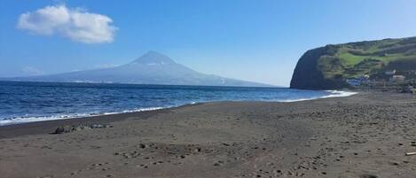 A beach with footprints leading to the water, a mountain in the background, and a cliff with buildings on the right.