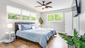 Un dormitorio con cama, ventilador de techo, televisor montado en la pared y ventanas amplias.