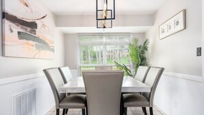 Un comedor con una mesa blanca, cuatro sillas, una lámpara colgante, un cuadro abstracto grande, una planta en maceta y una ventana con persianas.