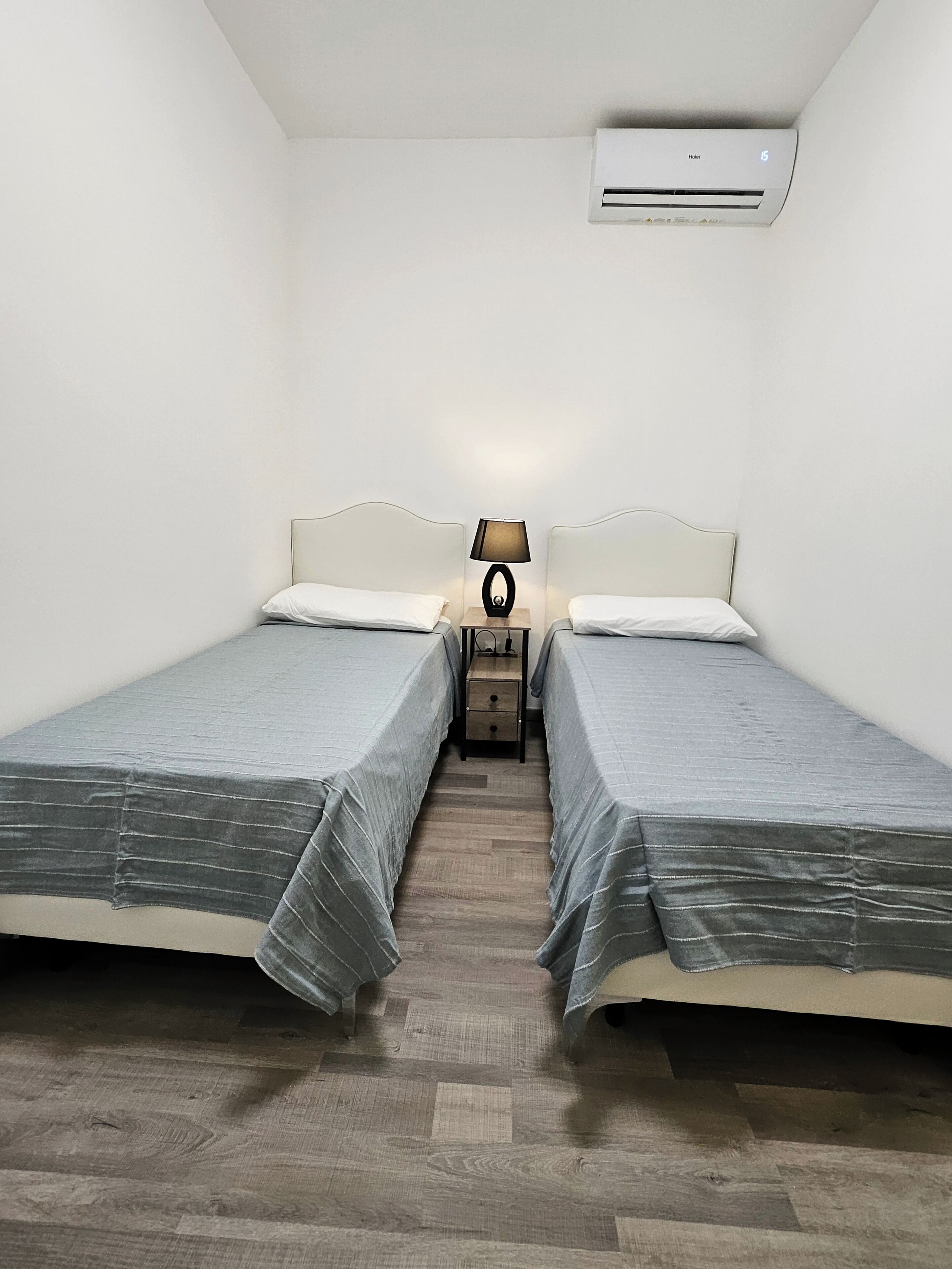 Une petite chambre propre, avec deux lits simples, une table de chevet avec une lampe et un appareil de climatisation fixé au mur.