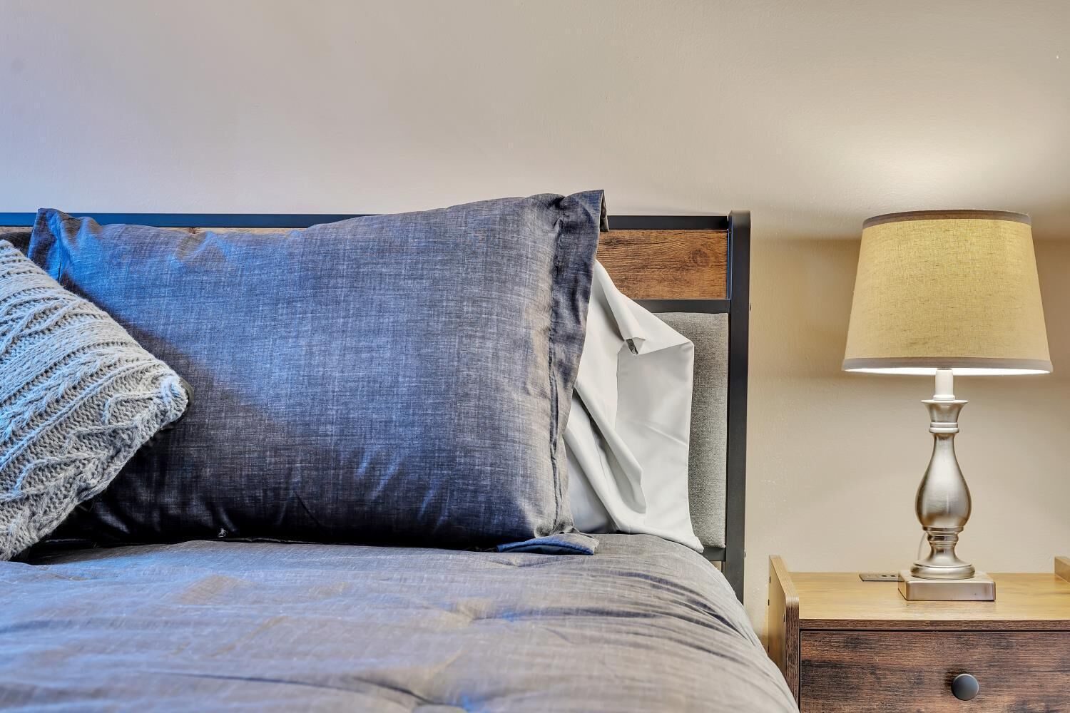 Una cama con una almohada gris y blanca, un plaid de punto y una mesita de noche con una lámpara.