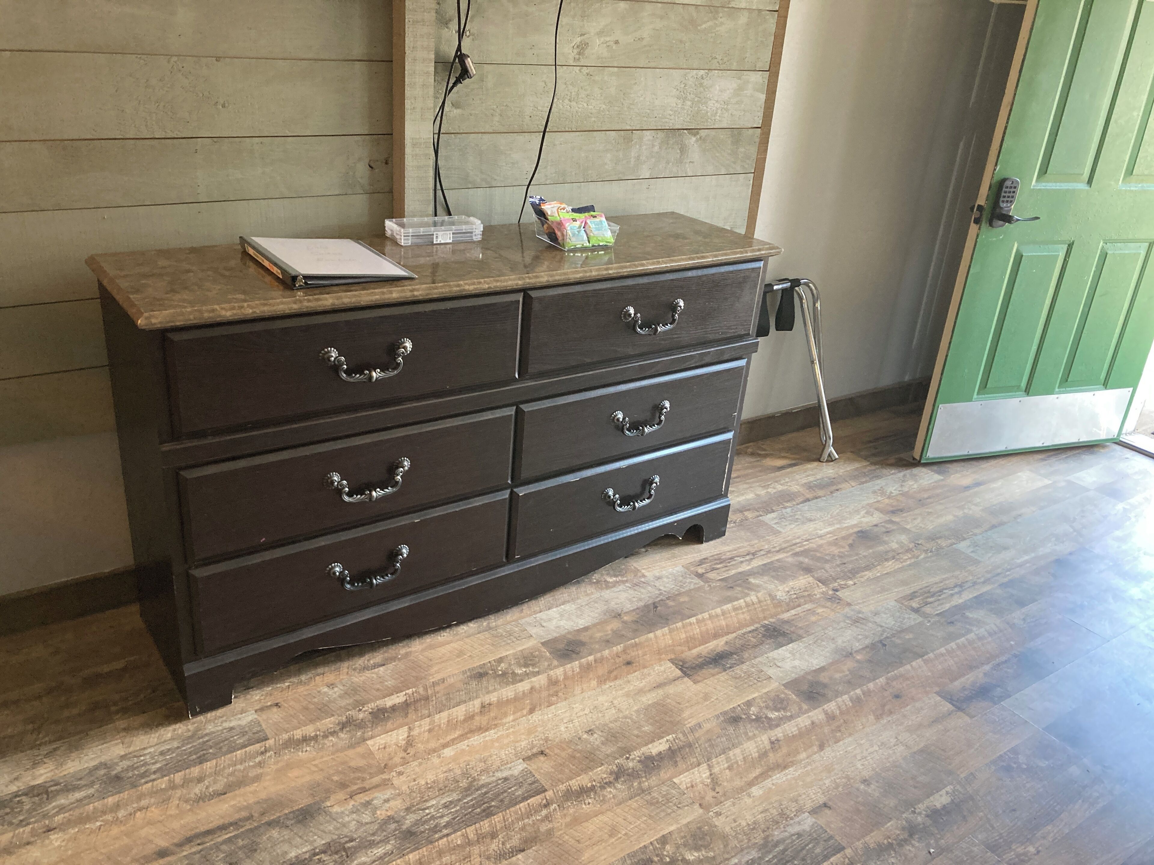 A wooden dresser with drawers and a top shelf, a green door, and a wooden floor.