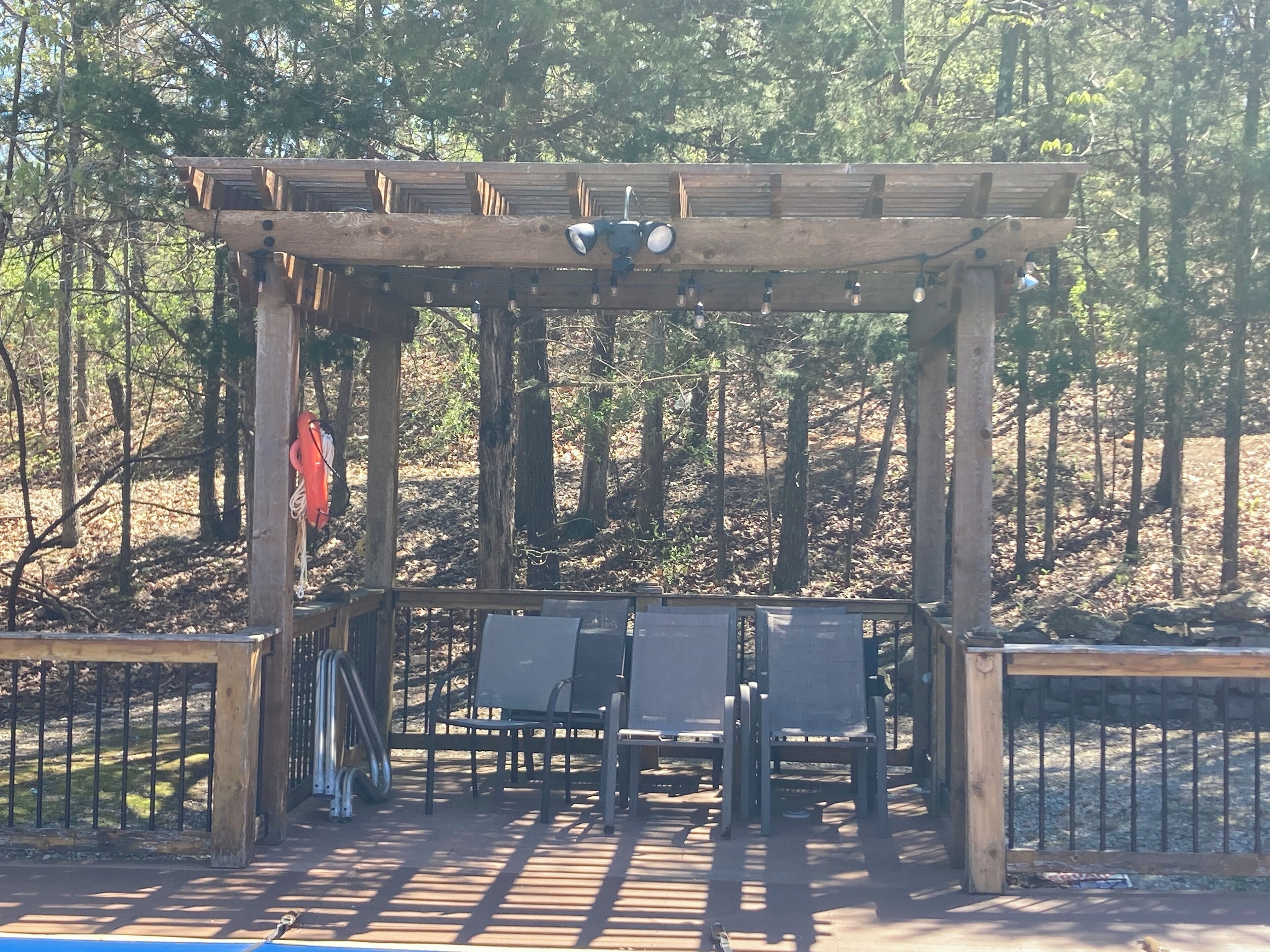 Een houten pergola met twee stoelen eronder, omgeven door een metalen hek, gelegen in een bebost gebied.