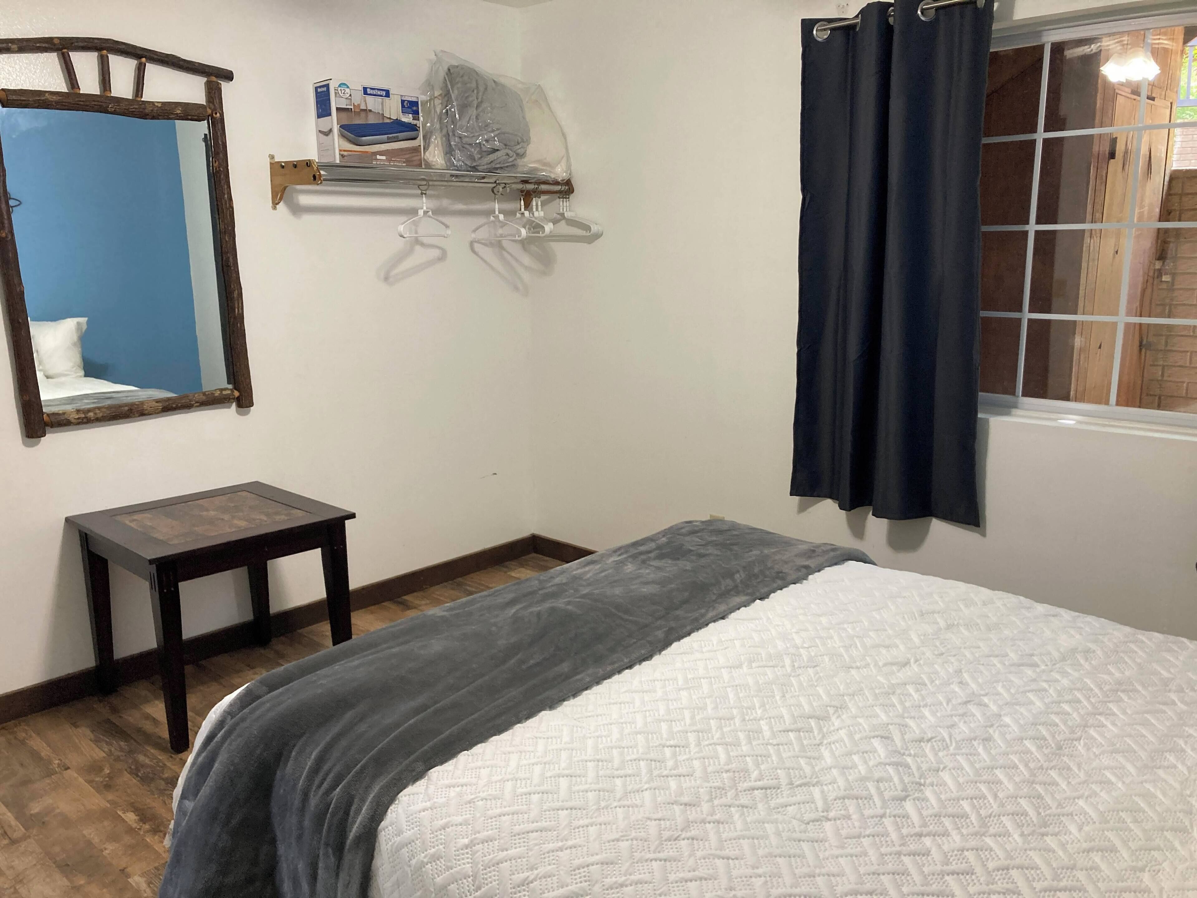 A bedroom with a bed, a bedside table, a mirror, a window with curtains, and a shelf with items.