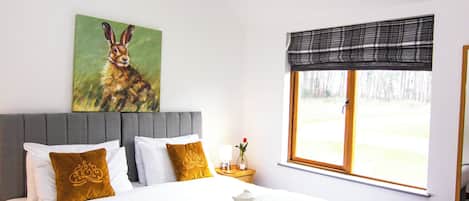 A neatly made bed with a green blanket, a painting of a rabbit on the wall, and a window with a plaid valance.