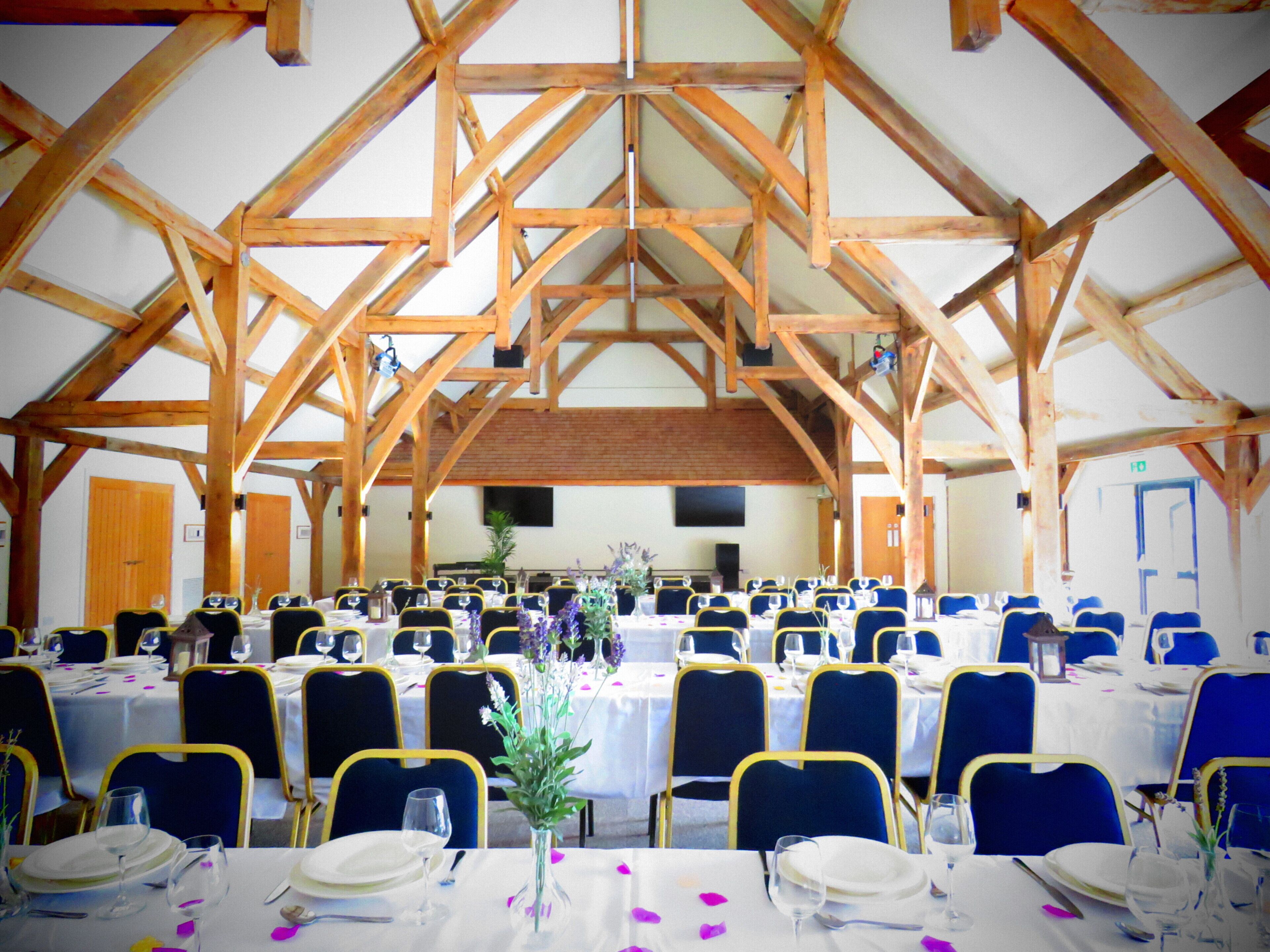 Een ruime zaal met hoge houten balken, tafels gedekt met witte tafelkleden, blauwe stoelen en bloemrijke decoraties.