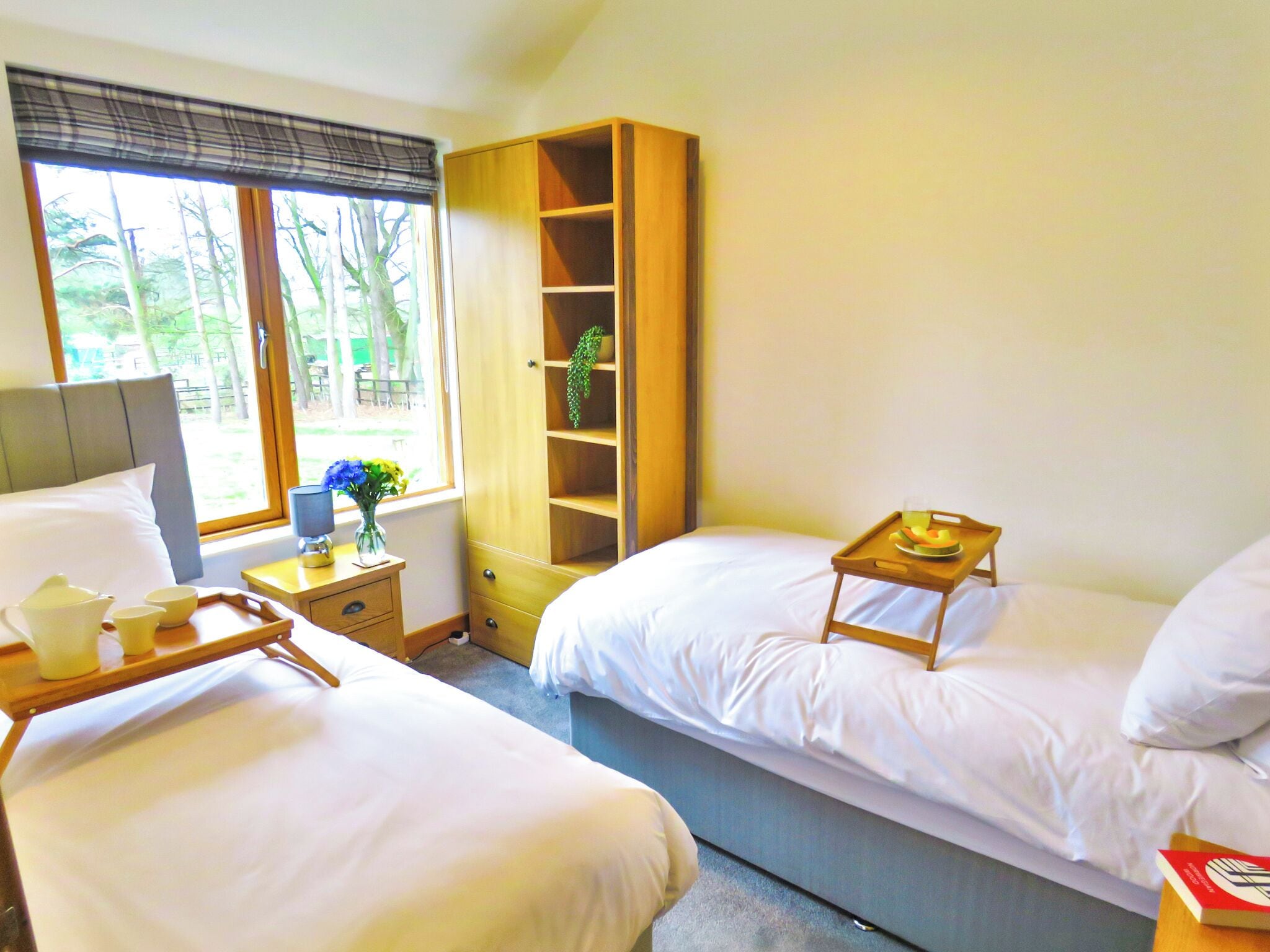 Ein Hotelzimmer mit zwei Betten, einem Holzschrank, einem Nachttisch mit Tablett und einem Fenster mit Blick auf Bäume.