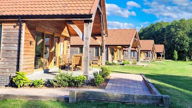 A row of wooden cabins with pitched roofs, each featuring a porch and a window. - Hay's Wood Retreat (King's Lynn)
