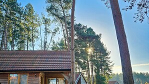 A wooden cabin with a tiled roof, a patio with a table and chairs, surrounded by a grassy area and tall trees. - Hay's Wood Retreat (King's Lynn)